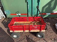 Front angle view of red wooden wagon with visible side rails, metal pull handle, and four pneumatic tires on gravel ground.