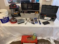 Overview of all items on a white table including Shop-Vac, tool boxes, booster cables, and hand tools