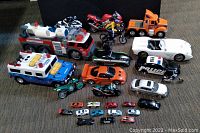 Wide view of all toy vehicles including the fire rescue truck, orange construction truck, police motorcycle, white convertible, various motorcycles, and small toy cars arranged on a carpeted floor.