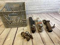 Lot showing three chrome trailer hitch balls, two receiver hitch adapters (one U-Haul PowerTow stainless steel, one rusted steel), and a gray plastic milk crate. Items are displayed on wood flooring.