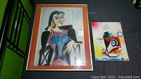 Photo shows two framed artworks placed on a floor: a larger cubist style portrait of a woman seated, and a smaller colorful abstract signed piece.