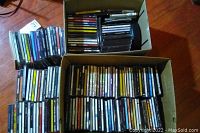 Two cardboard boxes filled with numerous CDs arranged primarily vertically inside the boxes and some stacked outside on the floor, showing a large assortment.
