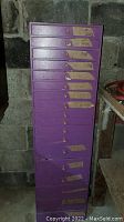 Front view of tall purple wooden chest of drawers showing many drawers with paper tags and several missing knobs.