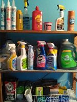 Three wooden shelves filled with assorted cleaning and household products including sprays, bottles, and sealants against a blue wall.