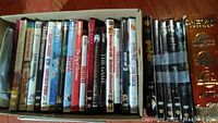 Photo of assorted DVDs in a box showing spines with various titles including The Joy of Christmas, Meet the Parents, Mamma Mia, and more, some with noticeable wear.