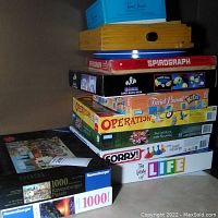 Stack of board games including Trivial Pursuit Kids, Spirograph, Card Magic Show, Operation, Sorry, The Game of Life, and puzzles stacked on the floor