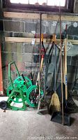 Various garden tools including hose reel, rakes, broom, post hole digger against a cinder block wall