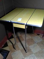 Yellow laminate kitchen table with two foldable sides, metal edging and metal legs in a room corner.
