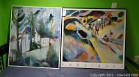 Two framed wall hangings on a green wall, left is oil painting with buildings and trees, right is Kandinsky abstract print