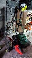 Photo showing Black & Decker electric hedge trimmer with sharp twin blade cutter, Ryobi gas-powered string trimmer with white fuel tank and green frame, and an orange and black extension cord all standing on a concrete floor in a garage.