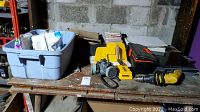 Wide view of tabletop showing tile cutter, gray plastic bin with supplies, Dewalt drill, black and orange toolbox, stacks of tiles, and other DIY items.