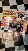 Photo showing a stack of multiple vintage advertising posters on a checkered floor, includes visible titles like 'The 3 Stooges Half Shot Shooters' and 'Robin Hood'. Posters show wear, hole punches on corners.