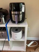 Gourmia air fryer on a shelf with Cuckoo rice cooker below it, both unplugged indoors near wall outlet.