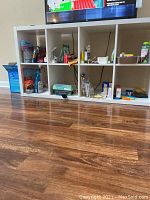 Photo showing the front view of white laminate cubicle shelf with 8 cubes filled with various household items, placed on wooden floor beneath a mounted TV.