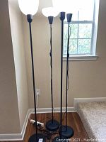 Four black metal floor lamps standing next to a wall and a window.