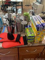 Overview of metal utensil containers with various kitchen tools, several boxes of foil and plastic wrap stacked, red silicone oven gloves on counter, and metal thermos.