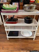 Photo of white metal shelving unit with various kitchen utensils including plastic trays, metal bowls, colander, pots, and pans