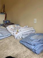 Stack of folded linens including light blue comforters, blankets, and white linens with animal prints on carpeted floor