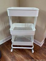 Front view of white metal rolling cart with three mesh baskets on wheels.
