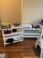 Two white metal shelving units, each with multiple metal mesh shelves, placed side by side against a wall with some kitchen and household items on and around them.