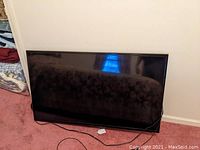 Front view of Insignia 48-inch LCD TV leaning against wall on carpet with power cable attached, no stand visible.