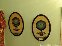 Photo of two oval framed antique hot air balloon prints hung side by side on a pale green wall.