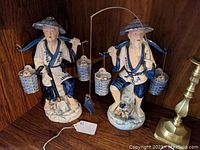 Pair of porcelain Chinese man and woman statues standing on a wooden shelf, each carrying two hanging baskets on a shoulder pole, painted predominantly blue and white with some gold highlights.