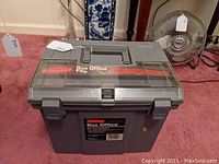 Gray Rubbermaid Box Office File TVR file box with clear hinged lid and carrying handle, shown closed on a carpeted floor beside a fan.