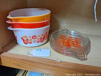 Three nested Pyrex Friendship pattern casseroles in white, orange, and yellow bases with spout handles. Glass lid with orange and red floral pattern to the right.