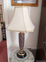Full view of the slender urn-shaped pottery lamp with connected beige paneled shade on lace tablecloth.