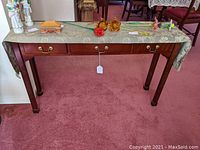 Front view of the mahogany colored wooden sofa table with three drawers and brass handles, covered partially by a floral table runner and topped with glass flowers and porcelain figurines.