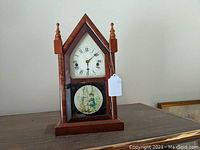 Full front view of wooden Gothic steeple clock on shelf, showing pointed top with finials and lower decorative tablet scene of children.