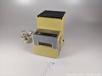 Vintage yellow Van Wyck electric ice crusher plugged in and standing on white surface, showing full unit front and top.