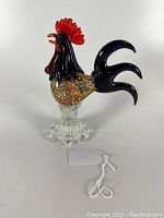 Front side view of the colorful art glass rooster figurine highlighting red comb and speckled body on a clear glass base.