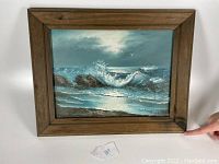 Framed oil painting showing ocean waves crashing rocks under moonlit cloudy sky with wooden frame.