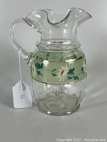 Clear blown glass lemonade pitcher with applied handle, hand-painted enamel floral band, and pontil mark on bottom, shown from a side angle with a white tag attached.