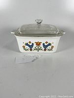 Front angle view of white Corning Ware casserole dish with colorful country festival pattern and clear glass lid.