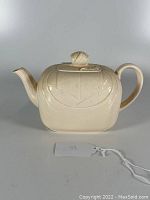 Front view of a beige ceramic teapot with embossed geometric Art Deco style design and shell-shaped knob on lid.