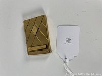 Closed gold-tone compact with criss-cross diagonal line pattern and a small attached lipstick holder on the front face, accompanied by a white price tag.