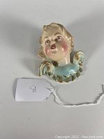 Front view of vintage ceramic angel bust with blond hair, rosy cheeks, and blue garment with gold leaf detailing.
