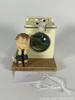Front view of the figurine showing Linus sitting on a towel beside a vintage yellow washing machine with Snoopy on top. The motto 'The best things in life are worth waiting for' is visible on the base.