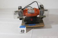 Front view of the bench grinder showing the entire tool with grinding stones and safety shields, mounted on a wooden board with a number 49 marker.