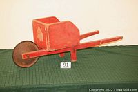 Full view of antique child's wooden wheelbarrow painted red, showing the tub, handles, and wheel on a green surface.