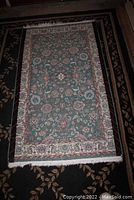 Full view of rectangular throw rug with blue background and floral geometric pattern, placed on a dark carpet with a leaf motif.