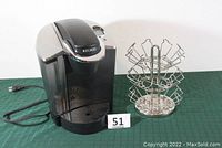 Keurig coffee maker and metal coffee pod carousel on green surface with lot number tag visible.