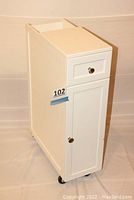 Front and side view of the white narrow rolling cabinet with drawer and door closed, showing wheels and overall shape