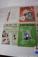 Four vintage sheet music covers laid flat: patriotic song with American flag, 'All Dressed Up With a Broken Heart' with male singer photo, 'Oh, But I Do' featuring black and white photos of performers, 'A Four Leaf Clover' in green with male trio photo.