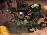 Wide angle view showing the green air compressor with attached pressure gauge, hoses coiled on top, motor, and a spare black plastic wheel.
