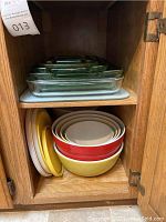 Anchor Hocking green and clear glass baking dishes stacked on the top shelf and melamine colored bowls with lids and a yellow bowl on the bottom shelf inside a wooden cabinet.