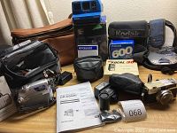 Photo showing assortment of cameras, camera bags, and accessories on table including JVC digital video camera with manual, Polaroid Impulse instant camera, Petri Color 35 camera, Focal 30 flash box, Seville 35mm camera, and various bags and small equipment.